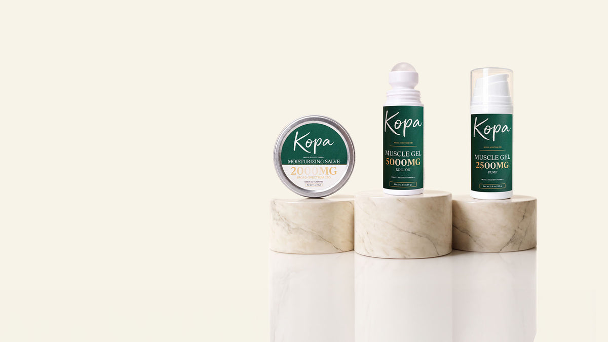 All three Kopa products on marble pedestals: the Moisturizing Salve tin, the Muscle Gel Roll-On 5000mg, and the Muscle Gel 2500mg pump